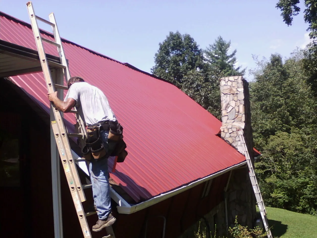 Roofer on a ladder beside a red metal roof and stone chimney — Soffit & Fascia Repair by Roofing Brothers in Middleport